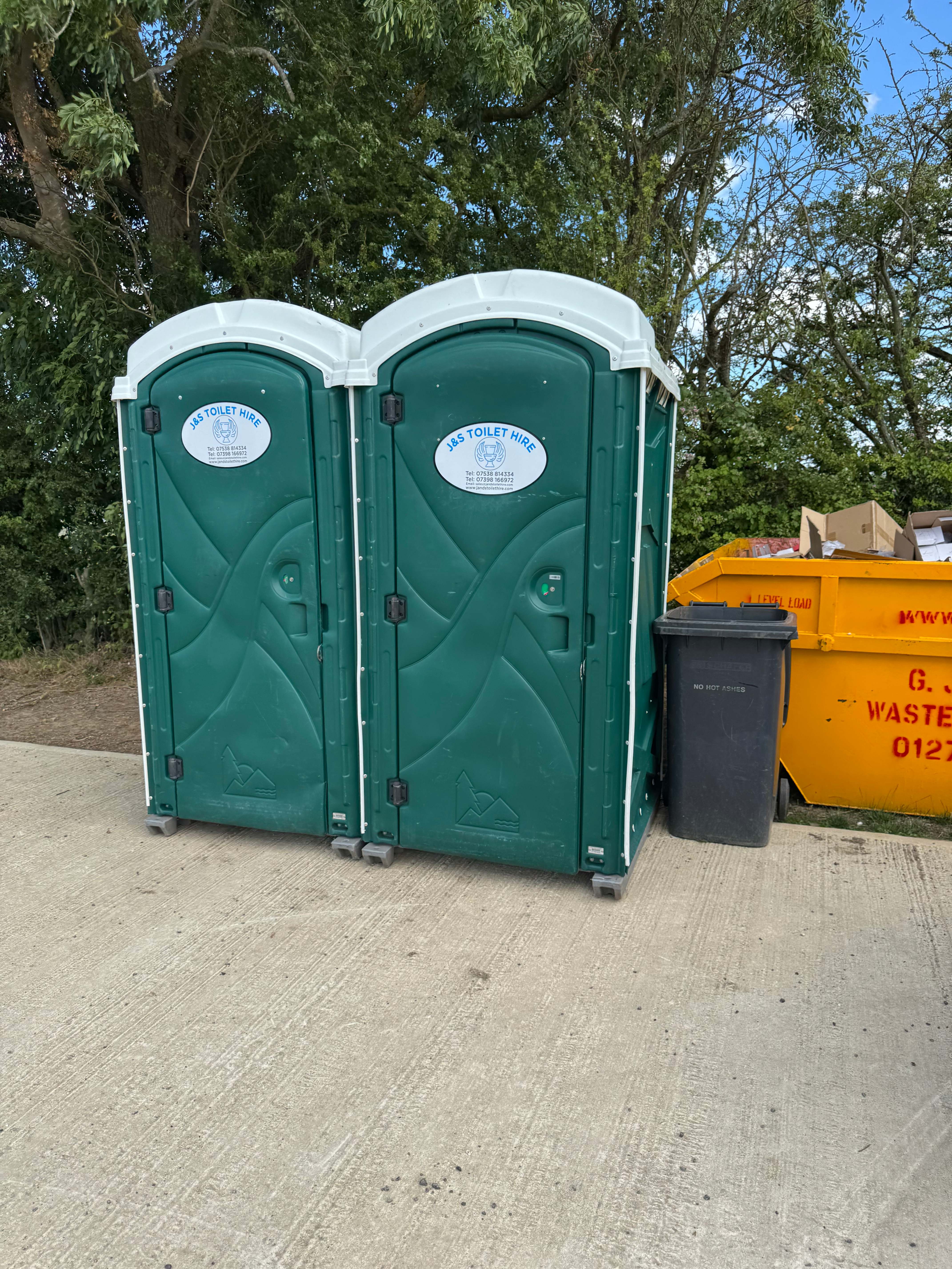 Two green J&S Toilet Hire portable toilets on concrete with trees in background